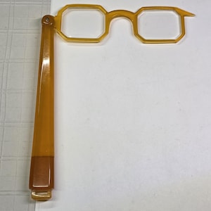 May include: A vintage-style, amber-colored eyeglass with a single arm. The glasses feature a rectangular frame design. The arm is attached with a hinge and has a rectangular end. The glasses are made of a translucent material.