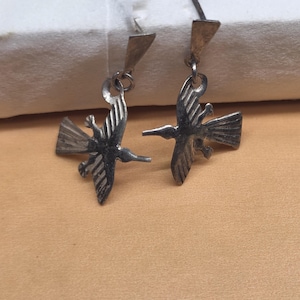 May include: A pair of silver-toned metal earrings featuring a bird-shaped pendant suspended from a triangular post. The bird design includes etched lines on the wings and tail, with a pointed beak and outstretched wings in a flying position.