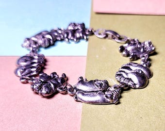 May include: A silver charm bracelet with a variety of charms, including pigs, hands, and other abstract shapes. The bracelet is on a light blue, pink, and green background.