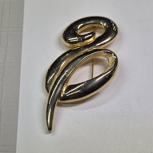 May include: A gold-toned brooch in a stylized, cursive letter "S" shape. The brooch has a pin on the back for attachment. The design features smooth, curved lines and a polished finish, reflecting light.