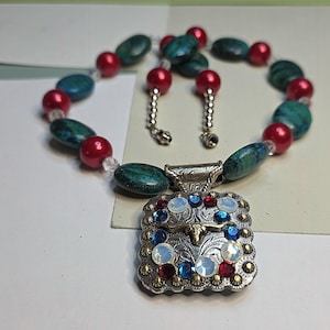 May include: A necklace and earring set featuring a silver pendant adorned with red, blue, and white gemstones. The necklace and earrings have red and teal beads. The jewelry set is on a white surface.