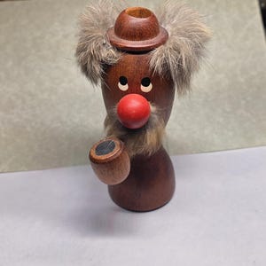 May include: A wooden clown figurine with a red nose, fur hair, and a small hat. The clown is holding a small wooden cup. The figurine is brown with black eyes and a fur mustache.