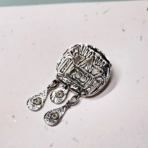 May include: A silver pendant with a geometric design and three dangling teardrop-shaped charms. The charms have a hammered texture and a small circle in the center.
