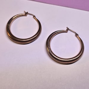 May include: Two rose gold hoop earrings. The earrings are round with a smooth, polished finish. The hoops are a medium size and have a simple, classic design. The earrings are displayed on a white and purple background.