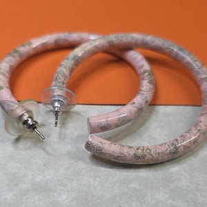 May include: A pair of pink and green patterned hoop earrings. The earrings have a marbled design with a post and back closure. The earrings are set against a neutral background with an orange backdrop.