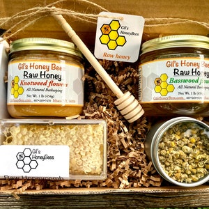 May include: A gift box filled with honey products from Gil's HoneyBees. The box contains two jars of raw honey, one with knotweed flowers and the other with basswood flowers, a honeycomb, a wooden honey dipper, and a tin of chamomile flowers.