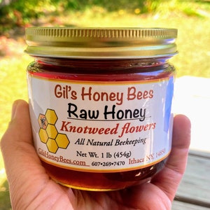 May include: A glass jar of raw honey with a gold lid. The label reads "Gil's Honey Bees Raw Honey Knotweed flowers All Natural Beekeeping Net Wt. 1 lb (454g) GilHoneyBees.com 607-269-7470 Ithaca NY. 14850"