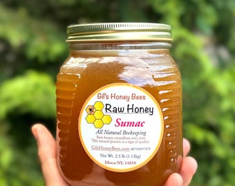 May include: A clear glass jar filled with amber-colored raw sumac honey, topped with a gold metal lid. The circular label features a honeycomb graphic, a bee icon, and the text Gil's Honey Bees, Raw Honey, Sumac, All Natural Beekeeping, Net Wt. 2.5 lb.