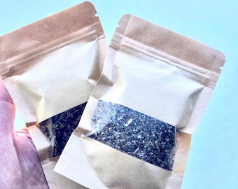 May include: Two brown paper pouches with clear plastic windows, each containing dried herbs. The pouches are held in a hand.