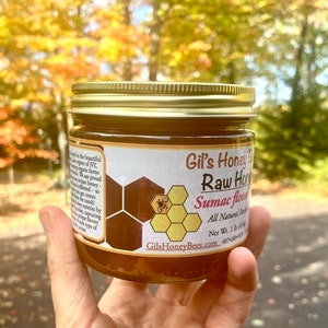 May include: A glass jar of raw honey with a gold lid. The label reads "Gil's Honey Bees Raw Honey Sumac Flower All Natural Beeswax Net Wt. 1 lb (454g) GilsHoneyBees.com 607-240-7099".