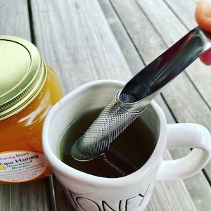 May include: A white ceramic mug with a handle, filled with a dark liquid and a stainless steel tea infuser. The infuser is being held by a hand, and a jar of honey is visible in the background.
