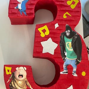 May include: Red number three piñata decorated with musical notes, stars, and cartoon characters. The piñata features characters from the movie "Sing" including a koala, gorilla, and pig. Perfect for a birthday party.