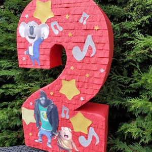 May include: Red felt number two piñata featuring a red, white, and blue koala, a black gorilla, and a pink pig from the film Sing. The piñata is decorated with gold stars and silver musical notes.