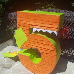 May include: Orange and green number five shaped pinata with a dragon design. The pinata has a green tail and a white mouth.