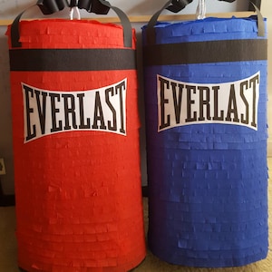 Puede incluir: Dos pinatas cilíndricas con forma de sacos de boxeo, una roja y otra azul. Cada una presenta una banda horizontal negra en la parte superior, una etiqueta blanca con el texto negro EVERLAST y un asa negra con un cordón blanco.