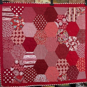 May include: A handmade quilt featuring a patchwork design of red hexagonal shapes. The quilt showcases various red patterns, including polka dots, floral prints, and stripes. The quilt is bordered with a red fabric and a white scalloped edge.
