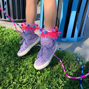 May include: Purple high-top sneakers with colorful pom-pom socks. The sneakers are on a bed of green grass with a multi-colored jump rope.
