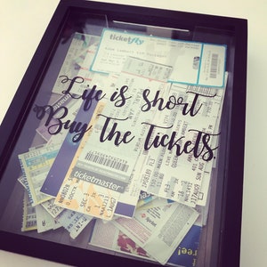 May include: A black framed shadow box filled with concert ticket stubs. The tickets are layered and overlapping. The text "Life is short, Buy the tickets." is written in black cursive lettering on the glass.
