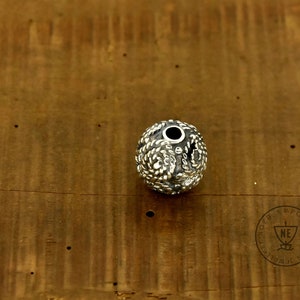 May include: A silver bead with a detailed, textured surface. The bead has a small hole in the center for stringing.
