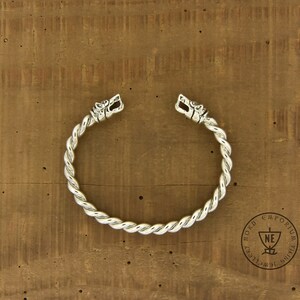 May include: A silver-colored Viking-style bracelet with a twisted rope design. The bracelet features detailed wolf head terminals. The jewelry is displayed on a wooden surface.