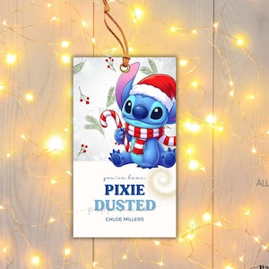 Puede incluir: Una etiqueta de regalo rectangular con un personaje alienígena azul que lleva un gorro de Papá Noel rojo y blanco y una bufanda a rayas a juego mientras sostiene un bastón de caramelo. El fondo tiene ramas de pino y bayas rojas.