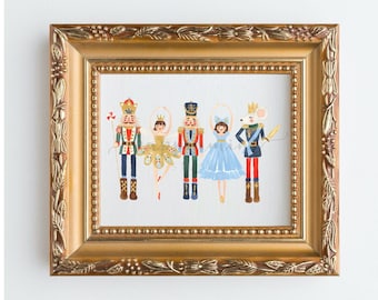 Puede incluir: Una ilustración enmarcada de un Cascanueces, una bailarina y un rey ratón. El Cascanueces lleva un uniforme rojo y dorado, la bailarina lleva un tutú dorado y blanco, y el rey ratón lleva un uniforme azul y dorado. La ilustración está sobre un fondo blanco.