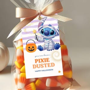Puede incluir: Una bolsa de plástico transparente llena de dulces de maíz naranjas, amarillos y blancos, atada con una cinta beige. Una etiqueta rectangular presenta un fondo de rayas azules, un personaje envuelto en vendas, una calabaza y el texto "you've been PIXIE DUSTED HAPPY HALLOWEEN!"