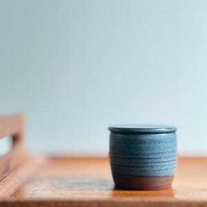 May include: A small, cylindrical ceramic jar with a lid, featuring a speckled blue-grey glaze on the upper portion and a smooth, unglazed reddish-brown base. The container has subtle horizontal ridges along the body and a flat, matching lid.