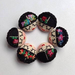 May include: Five embroidered fabric buttons with black hair, white faces, and colourful floral designs. The buttons are arranged in a circle.