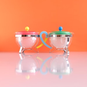 Pode incluir: Duas chaleiras de vidro transparente com tampas e alças coloridas. Uma tem uma tampa rosa com um botão amarelo, a outra tem uma tampa verde com um botão azul. As alças são azuis e amarelas. As chaleiras estão sobre uma superfície branca contra um fundo laranja.