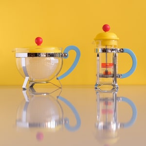 Könnte beinhalten: Zwei Glas-Kaffeepressen mit gelben Deckeln und blauen Griffen vor gelbem Hintergrund. Die Pressen haben einen silbernen Rahmen und einen roten Knopf. Eine ist teekannenförmig, die andere eine French Press.