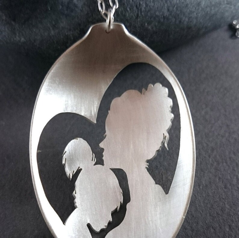 Pu&ograve; includere: Una collana a cucchiaio d'argento con un design a forma di cuore ritagliato che rappresenta una madre e un bambino.