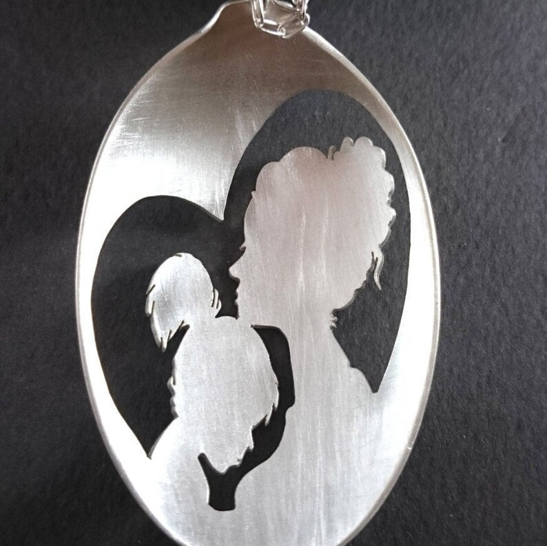 Pu&ograve; includere: Un cucchiaio d'argento con un ritaglio a forma di cuore che presenta una silhouette di una madre e un bambino. Il cucchiaio &egrave; un ciondolo e ha una catena d'argento.