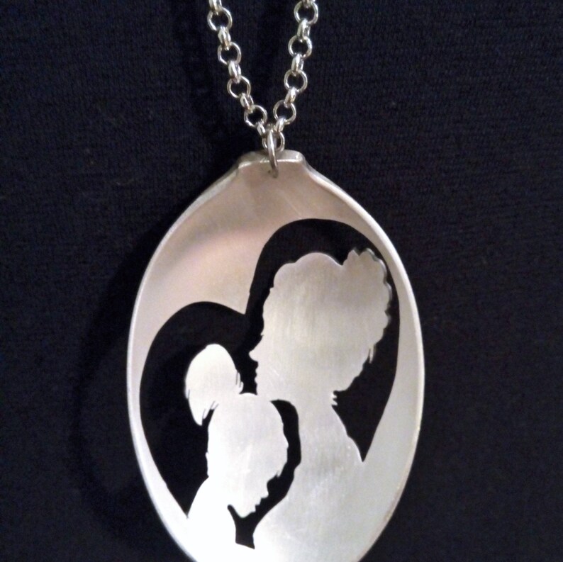 Pu&ograve; includere: Una collana a cucchiaio d'argento con un design a forma di cuore con due figure ritagliate. Le figure sono una madre e un figlio.
