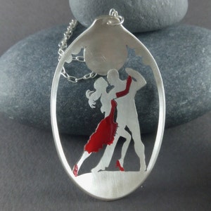 Puede incluir: Un collar con un colgante de plata que presenta una silueta recortada de una pareja bailando tango. La mujer lleva un vestido rojo y el hombre lleva un traje. El colgante tiene forma ovalada y tiene una cadena unida a él.