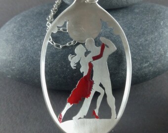 May include: A silver pendant necklace featuring a cutout silhouette of a couple dancing the tango. The woman is wearing a red dress and the man is wearing a suit. The pendant is shaped like an oval and has a chain attached to it.
