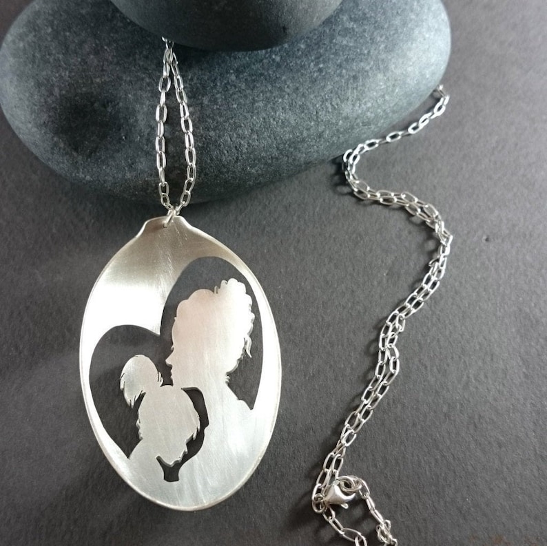 Pu&ograve; includere: Una collana d'argento con un ciondolo a forma di cucchiaio. Il ciondolo presenta un design ritagliato di due persone, una madre e un figlio, in piedi uno accanto all'altro all'interno di un cuore.