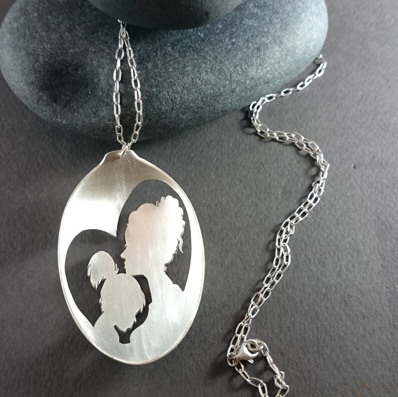 Pu&ograve; includere: Una collana d'argento con un ciondolo a forma di cucchiaio. Il ciondolo presenta un design ritagliato di una madre e un figlio che si abbracciano.