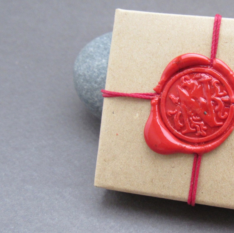 Pu&ograve; includere: Una piccola scatola di cartone marrone legata con uno spago rosso e sigillata con un sigillo di cera rossa. Il sigillo di cera ha un disegno di un uccello stilizzato.