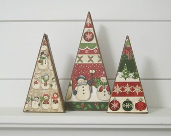 May include: Three wooden Christmas tree decorations. The trees are triangular and decorated with festive patterns, including snowmen, snowflakes, and ornaments. The trees have brown edges and are arranged on a white surface.