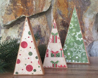 May include: Three decorative Christmas tree-shaped ornaments. The trees have a triangular shape with a wooden frame. The patterns include red and green polka dots, gift boxes, and snowflakes. The background is a stone wall.