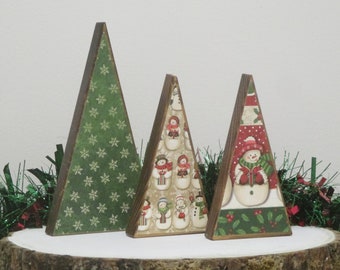 May include: Three wooden Christmas tree decorations. The trees are triangular, with a rustic wood edge. One tree has a green snowflake pattern, and the other two feature snowman designs. The trees are displayed on a white surface.