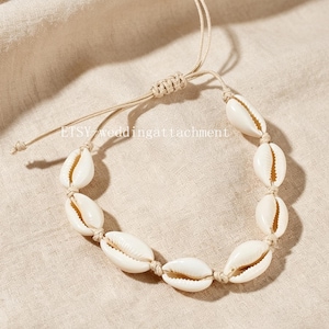 Peut inclure: Un bracelet composé d'une série de coquillages cauris blancs enfilés sur un cordon beige clair. Le bracelet comprend un mécanisme de fermeture à nœud coulissant réglable et affiche le texte ETSY-weddingattachment au centre.