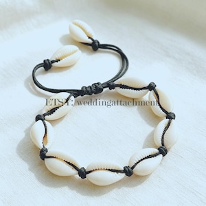 May include: A bracelet featuring white cowrie shells strung on a black cord. The design includes an adjustable sliding knot closure with two additional cowrie shells at the ends of the cord. The text ETSY: weddingattachment is visible.