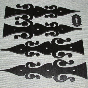 May include: Four black wooden decorative hinges with intricate scrollwork designs. The hinges are arranged in a vertical stack, with the top hinge slightly offset to the right. Each hinge has two holes for attaching to a surface.