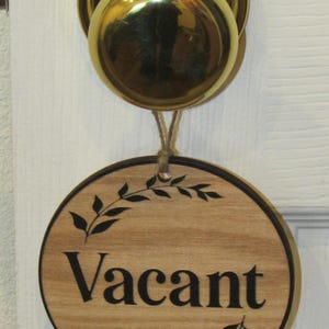 May include: A round wooden sign hanging from a polished brass doorknob. The sign features a light wood grain finish with the word Vacant printed in black, centered between two black leaf branch illustrations at the top and bottom of the text.