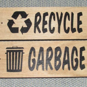 May include: Two wooden signs with black text and symbols. The top sign says "RECYCLE" with a recycle symbol. The bottom sign says "GARBAGE" with a trash can symbol.