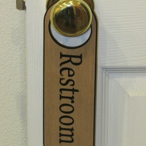 May include: A wooden door hanger with the word "Restroom" printed in black lettering. The hanger is attached to a gold door knob.