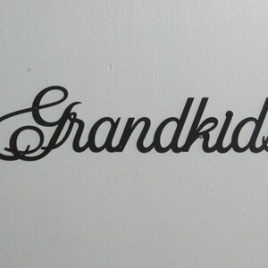 May include: Black metal sign that reads "Grandkids" in a cursive font. The sign is mounted on a light blue wall. The letters are connected and have a decorative style.
