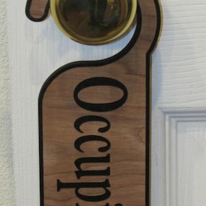 May include: A wooden door hanger with a light brown wood grain finish and a black border. The word "Occupied" is printed in large, bold, black capital letters vertically down the center of the hanger, which is hooked over a round brass doorknob.